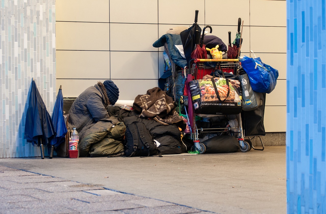 A homeless person crouching in a station with all their belongings around them, including bags and umbrellas in shopping cart.